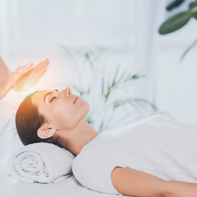 peaceful young woman with closed eyes receiving reiki treatment on head